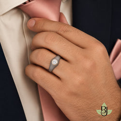 A high-resolution commercial product photograph capturing a close-up view of a man's right hand adjusting a dusty rose pink silk tie against a cream dress shirt and navy blue suit jacket. The man's hand, featuring a warm tan skin tone with highly visible pores, fine dark hair on the back and wrist, and short clean fingernails, is the central focal point, positioned diagonally across the frame from bottom right to top left. On the ring finger, he wears a uploaded product, The clothing consists of a dark navy blue suit blazer with a visible fine woven texture, a soft cream or off-white dress shirt with a subtle sheen and visible stitching along the placket, and a satin-finish dusty rose tie that is being gently grasped by the thumb and fingers. A matching dusty rose pocket square is tucked into the suit's breast pocket on the right side, adding a cohesive color element. The lighting is soft and diffused studio lighting, likely from a large softbox positioned to the upper left, which highlights the texture of the skin, the weave of the fabric, and the sparkle of the diamond while casting soft, natural shadows between the fingers. The depth of field is shallow, keeping the hand and ring in sharp focus while the navy suit background is slightly blurred, drawing the eye to the jewelry. A faint, semi-transparent watermark text reading "Bilipatra Jewels" is superimposed across the center of the hand in a grey serif font. In the bottom right corner, a brand logo displays a stylized "BJ" monogram in black within a circular design surrounded by light sage green leaves, with the text "Bilipatra Jewels" printed below in a small, dark font. The composition is intimate and elegant, emphasizing luxury and fine detail, with a sophisticated color palette dominated by deep navy, soft cream, muted pink, warm skin tones, and metallic grey and gold accents. The image quality is ultra-high definition, showcasing 8k level detail in the fabric textures and skin pores. output put image quality 2100 x 2100 pixels.