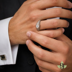 A close-up, high-resolution photograph of a man's hands clasped together in a formal setting, showcasing a uploaded product on the ring finger. The subject is wearing a dark black suit jacket and a crisp white dress shirt with French cuffs. On the left cuff, visible on the left side of the frame, is a rectangular cufflink featuring a silver metallic border and a dark, possibly black onyx or enamel center insert. The man's skin is tanned with realistic texture, showing visible pores, fine lines on the knuckles, and fine hairs on the forearm near the wrist. The left hand is positioned on top, palm facing down, displaying a distinctive ring on the ring finger. The right hand is positioned below, reaching up to gently grasp the left wrist, with the thumb pressing against the inner forearm and the fingers curling naturally over the back of the left hand and wrist. The fingernails are clean, short, and neatly trimmed. The lighting is soft and diffused, typical of professional studio product photography, creating gentle highlights on the knuckles, the curve of the ring band, and the facets of the diamond, while casting soft, natural shadows between the fingers. The background consists of the dark, out-of-focus fabric of the black suit jacket, with a single round black button visible near the bottom center. The image utilizes a shallow depth of field, keeping the ring and the central interaction of the hands in sharp focus while the cufflink and the background suit are slightly blurred, emphasizing the jewelry. The overall mood is elegant, sophisticated, and masculine, with a color palette dominated by neutral skin tones, stark white, deep black, and the metallic accents of silver, grey, and rose gold. The shot is captured with a macro lens, likely around 100mm, at a wide aperture to isolate the subject, resulting in an ultra-detailed, 8k quality image with sharp focus on the ring's texture and the skin's details. output put image quality 2100 x 2100 pixels.