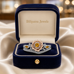 A realistic high-resolution product photograph captures an open navy blue velvet jewelry box positioned on flowing off-white satin fabric with soft natural folds and delicate sheen, the box featuring a polished gold metal accent lining the lid rim and a small round gold clasp button centered on the front lower edge. Inside the lid, elegant gold serif lettering reads "Bilipatra Jewels" against a smooth cream interior lining, while the lower compartment holds a uploaded ring. Soft diffused studio lighting from above and slightly to the right creates gentle highlights on the diamond facets, gold trim, and velvet texture while producing subtle shadows that emphasize depth and three-dimensional form. The background is heavily blurred with warm creamy bokeh orbs suggesting distant jewelry display cases under soft ambient lighting. The image is shot on a full-frame DSLR such as a Canon EOS R5 with an 85mm f/1.4 macro lens at a low aperture for razor-sharp focus on the ring and box against a creamy shallow depth of field, using a moderate ISO with clean color grading that delivers warm luxurious tones, subtle contrast, and a high-end commercial aesthetic reminiscent of premium jewelry catalog photography, ultra-detailed textures visible on the velvet surface, satin drape, metallic gold edges, and the gemstone's precise cut, all composed with the open box filling most of the frame and the ring positioned as the clear focal point in elegant symmetry. Output image quality must be exactly 2100 x 2100 pixels.