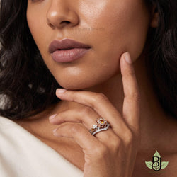 A realistic close-up photograph of a woman with warm olive skin and dark wavy hair, captured in a luxurious jewelry portrait style. The frame focuses tightly on the lower half of her face and her raised left hand, which is positioned gracefully near her jawline with fingers gently relaxed and slightly curved, the index finger extended upward in a delicate pose. Uploaded Ring rests on her ring finger, Her full lips are softly defined with a muted mauve-nude lipstick, her nose and chin are softly lit, and her skin displays a natural dewy glow with smooth texture and gentle highlights along the cheekbones. She wears an off-white soft fabric garment that drapes loosely across her shoulder and chest, creating subtle folds in the foreground. Soft diffused studio lighting illuminates the scene from the front and slightly above, producing even skin tones, delicate shadows under the fingers, and sparkling highlights on the diamond and gold metal. The image uses a shallow depth of field with the ring and hand rendered in crisp sharp focus while the background hair and fabric gently blur into a creamy bokeh. Precise colors include warm beige skin tones, and muted off-white fabric against the dark hair framing the composition. The ring sits slightly off-center in the lower right of the frame, balanced by the partial view of her face occupying the upper left and center. Professional studio photography with natural color grading, fine detail on skin pores and metal surfaces, elegant intimate mood, ultra high resolution, photorealistic clarity. output image quality 2100 x 2100 pixels.