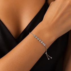 A high-resolution, macro product photograph capturing a close-up view of a woman's left wrist and forearm adorned with a uploaded bracelet. The arm is positioned diagonally across the frame, extending from the bottom left corner towards the top right, creating a dynamic composition against a dark background. The skin tone is a warm, light tan with highly detailed texture, revealing natural pores, fine lines, and a subtle, healthy sheen, particularly on the upper right side of the wrist where the light hits. The subject is wearing a black garment, likely a silk camisole or tank top with a V-neckline, which is visible in the upper left corner, showing a blurred section of the chest and collarbone area. The black fabric provides a high-contrast background that makes the jewelry pop. reflecting the studio lighting with bright white highlights. The lighting is soft and directional, originating from the upper right, casting gentle shadows and highlighting the contours of the arm and the brilliance of the stones. The photography utilizes a shallow depth of field, likely shot with a 100mm macro lens at an aperture of f/2.8 to f/4, ensuring the bracelet and the immediate skin texture are in sharp focus while the background elements and the lower forearm blur into a soft bokeh. The color palette is dominated by warm beige and peach skin tones, the bright silver and white of the diamonds and metal, and the deep, matte black of the clothing. The overall aesthetic is elegant, luxurious, and intimate, typical of high-end jewelry advertising or e-commerce product photography, rendered with ultra-high detail, realistic skin rendering, and a polished commercial finish. output image quality 2100 x 2100 pixels.