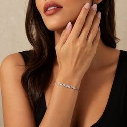 high-resolution fashion photograph capturing the upper torso and lower face of a woman with warm, light-medium skin tone, serving as an elegant product shot for delicate jewelry. The composition is tightly cropped, excluding the eyes and forehead, focusing on the graceful interaction between her hand and face. She has long, dark brown hair that falls over her left shoulder, visible on the left edge of the frame. Her lower face features full lips painted in a matte dusty rose or terracotta shade, slightly parted to show a hint of teeth, and a defined, smooth jawline. Her right hand is raised in a gentle, contemplative pose, with the palm facing her face and the tips of her index, middle, and ring fingers resting against her cheek and jaw. The nails are long, almond-shaped, and painted a soft, translucent pale pink nude color. On her right wrist, wear a uploaded bracelet. She is wearing a simple black sleeveless top with a wide, deep scoop neckline, likely a tank top or camisole, made of a matte, soft fabric that absorbs light and provides a strong contrast to her warm skin tone and the gold jewelry. The lighting is soft and diffused studio lighting, likely from a large softbox positioned to the front-right, which evenly illuminates her skin, revealing natural texture such as pores, fine lines, and vellus hair on her forearm, while casting soft, natural shadows under her chin and along her collarbone. The background is a plain, neutral beige or off-white wall with a very subtle plaster texture, kept slightly out of focus to draw attention to the subject. The image utilizes a shallow depth of field, characteristic of an 85mm portrait lens at f/1.8, keeping the bracelet and the hand in sharp focus while the background and the upper part of the face are slightly softer. The color palette is warm and neutral, dominated by skin tones, deep black, warm gold, and beige, creating an elegant, sophisticated, and minimalist aesthetic suitable for high-end fashion or jewelry advertising, rendered with 8k ultra-detailed quality and natural color grading. output image quality 2100 x 2100 pixels.
