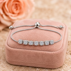A high-resolution macro product photograph of an elegant uploaded bracelet resting on a soft, blush-pink velvet jewelry box. The jewelry box is a square, cushioned case with a suede-like texture in a muted dusty rose or peach color, providing a soft, matte contrast to the shiny metal. The box rests on a textured, mottled beige and tan stone surface resembling marble or travertine, adding an organic, earthy base to the composition. In the upper left corner, a large peach-colored rose is visible but heavily blurred due to a shallow depth of field, creating a soft, romantic bokeh effect. The lighting is soft and diffused studio lighting, typical of high-end jewelry photography, casting gentle shadows beneath the box and creating bright specular highlights on the metal curves and the faceted stones. The depth of field is very shallow, likely shot with a 100mm macro lens at f/2.8, keeping the central heart motif and the infinity loop in sharp focus while the background rose and the rear of the box fade into a smooth blur. The color palette is warm and elegant, dominated by cool silver and white tones against warm blush pink, peach, and beige backgrounds. The overall mood is luxurious, delicate, and romantic, emphasizing the sparkle and craftsmanship of the jewelry. The composition is centered and balanced, with the bracelet forming a horizontal oval shape that draws the eye to the central heart. The image is ultra-detailed, 8k resolution, capturing every reflection and texture with photographic accuracy. output image quality 2100 x 2100 pixels.